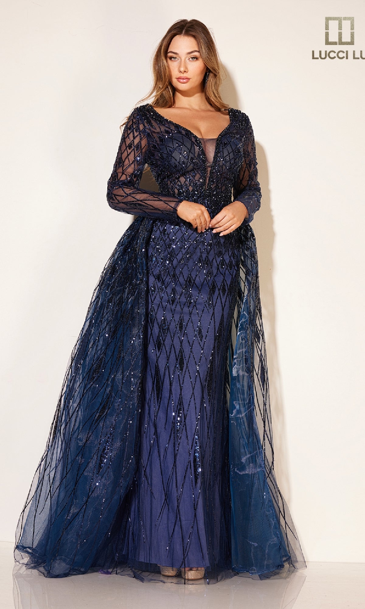 A woman wears the Lucci Lu C8095 long formal dress—a dark blue sequin gown with sheer sleeves and a tulle overskirt—standing against a plain light background. The Lucci Lu logo is shown in the top right corner.